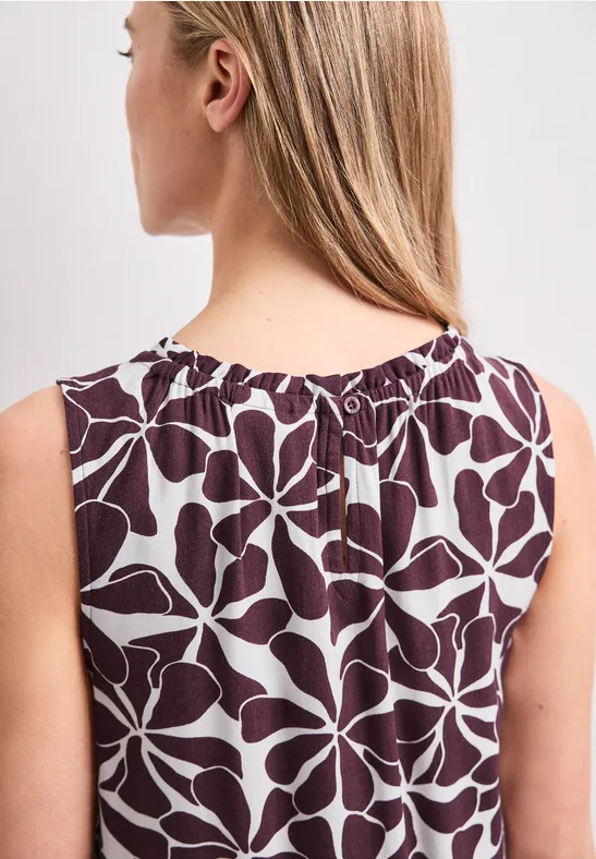 Rugaanzicht van een vrouw met lang blond haar in een mouwloze wijnrode top met lichtblauwe bladprint.