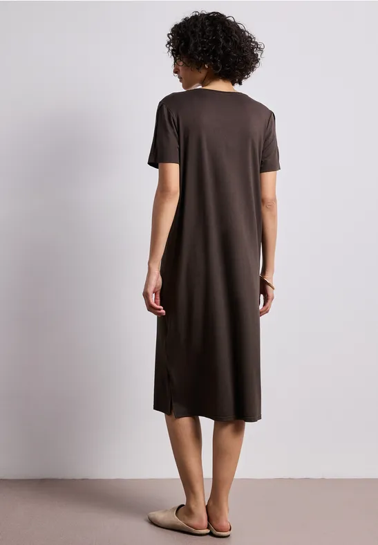 Eine Frau mit kurzem lockigem Haar steht vor neutralem Hintergrund, trägt ein schlichtes braunes Midikleid und Sandalen.