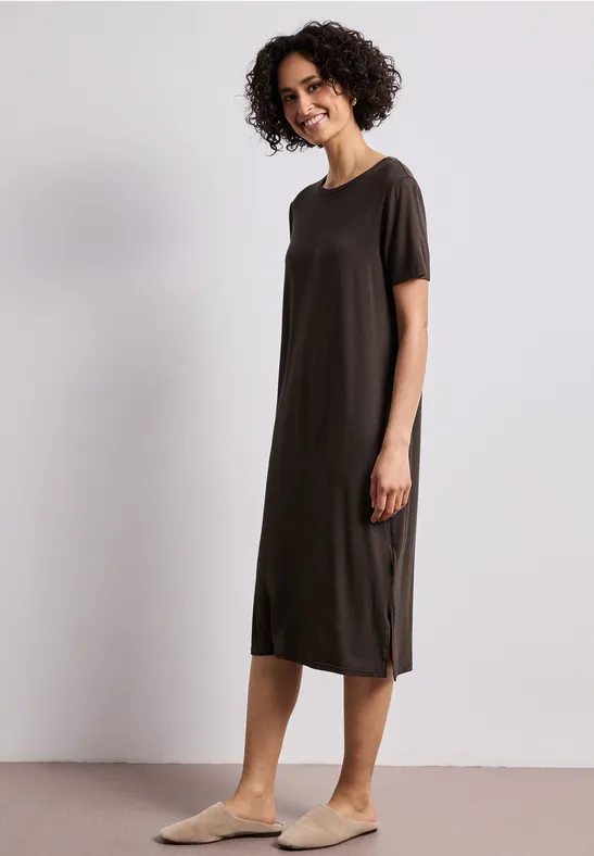 Eine Frau trägt ein schlichtes dunkelbraunes Kurzarm-Midikleid und bequeme Hausschuhe vor neutralem Hintergrund.