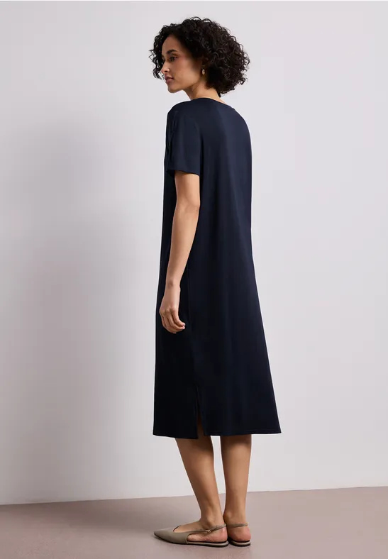 Frau in schlichtem, knielangem Marineblau-Kleid mit kurzem Arm und Rückenschlitz, flache Sandalen.
