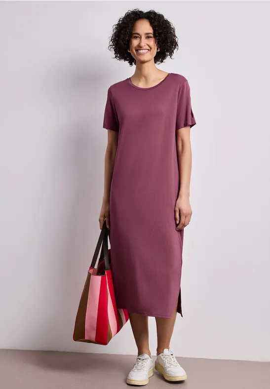 Person in lila Midi-T-Shirt-Kleid, weiße Sneaker und mehrfarbige Tragetasche.