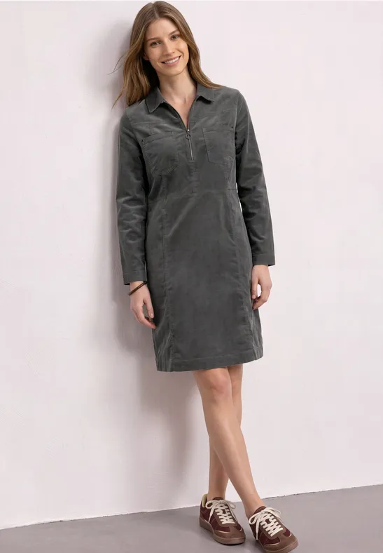 Vrouw in grijze corduroy jurk met kraag en lange mouwen en bruin-witte veterschoenen voor een witte muur.