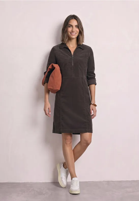 Frau in dunkelbraunem Cordkleid mit halbem Reißverschluss, weißen Sneakern und orangem Clutch.