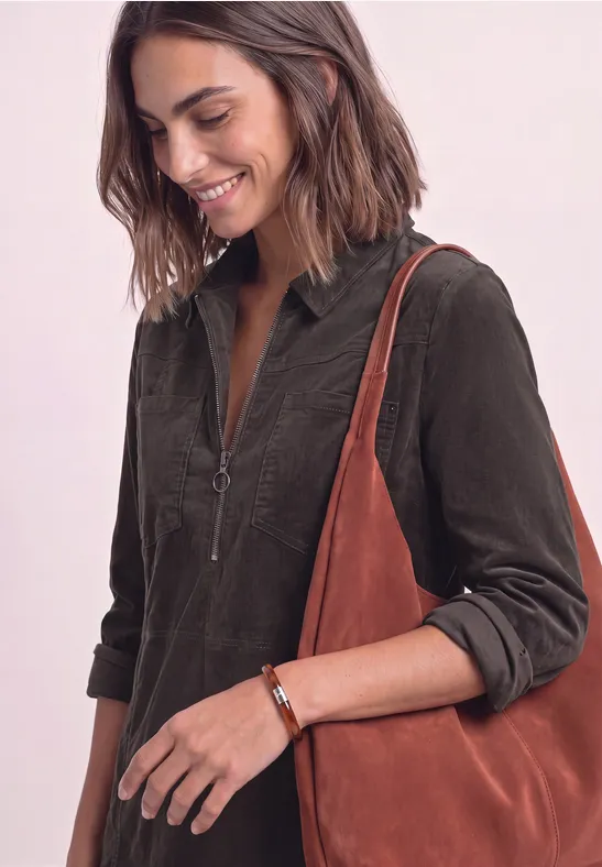 Eine lächelnde Frau trägt ein dunkelgrünes Cordkleid mit Reißverschluss und eine große braune Wildledertasche.