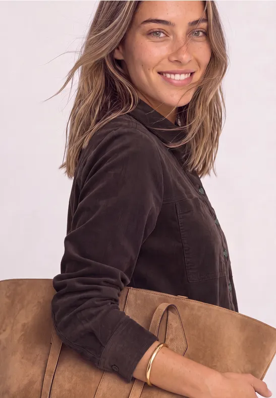 Lächelnde Frau mit brauner Wildledertasche und dunkler Jacke schaut in die Kamera.