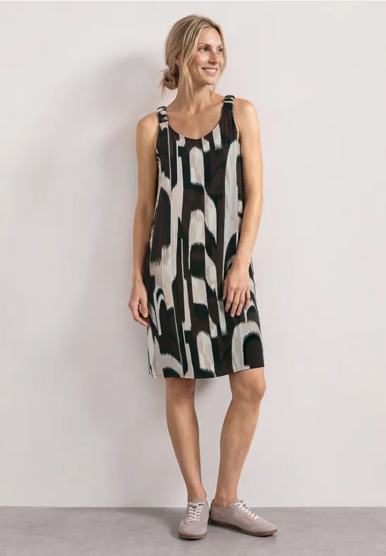 Femme portant une robe sans manches à motif noir et blanc et des chaussures grises devant un mur clair.