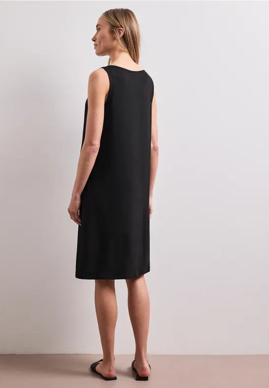 Frau von hinten in ärmellosem, knielangem schwarzen Kleid und flachen Sandalen vor neutralem Hintergrund.