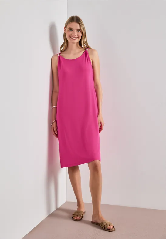 Vrouw poseert in mouwloze, knielange fuchsia jurk met sandalen voor een effen achtergrond.