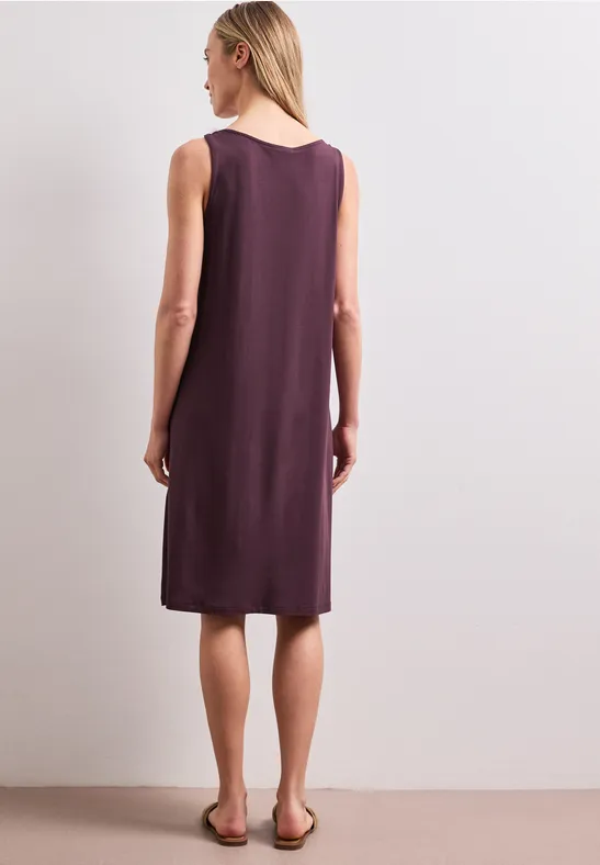 Rückenansicht einer Frau in ärmellosem, knieumspielendem, dunkellila Kleid und flachen Sandalen.