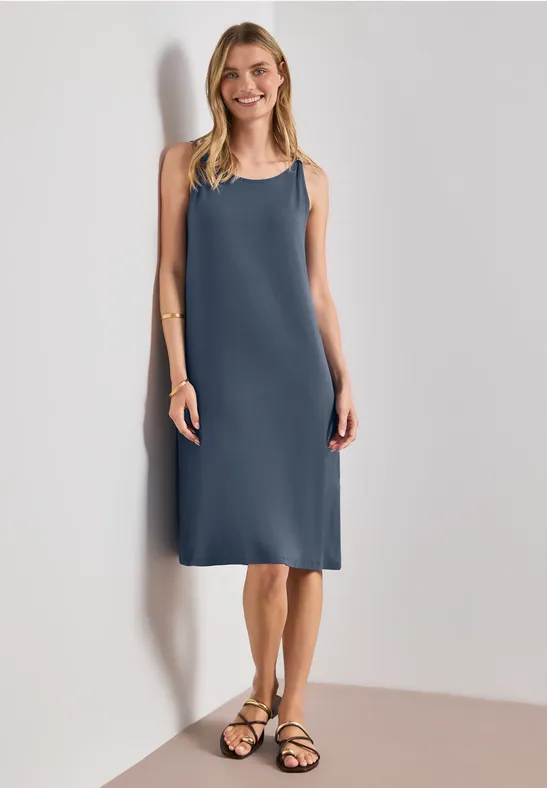 Frau in ärmellosem knielangem blauem Kleid und schwarzen Sandalen vor hellem Hintergrund.