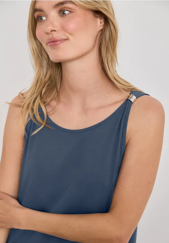 Eine junge Frau mit hellblonden Haaren und blauem Trägertop, leicht seitlich im Studio posiert.