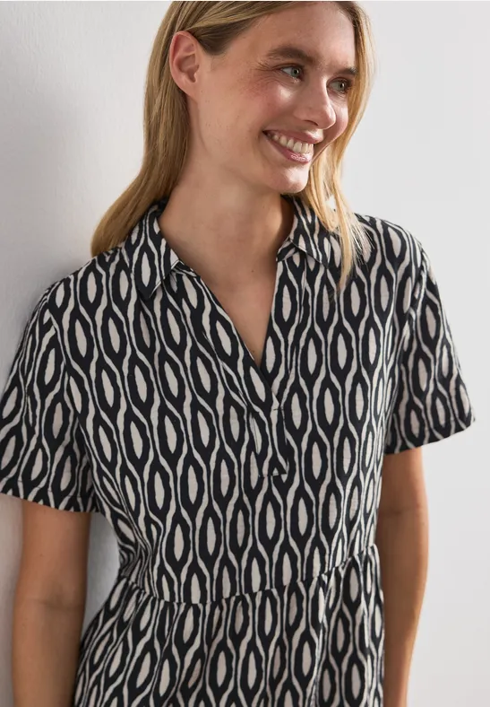Frau im schwarz-weißen, kurzärmeligen Kleid mit geometrischem Muster vor weißer Wand.