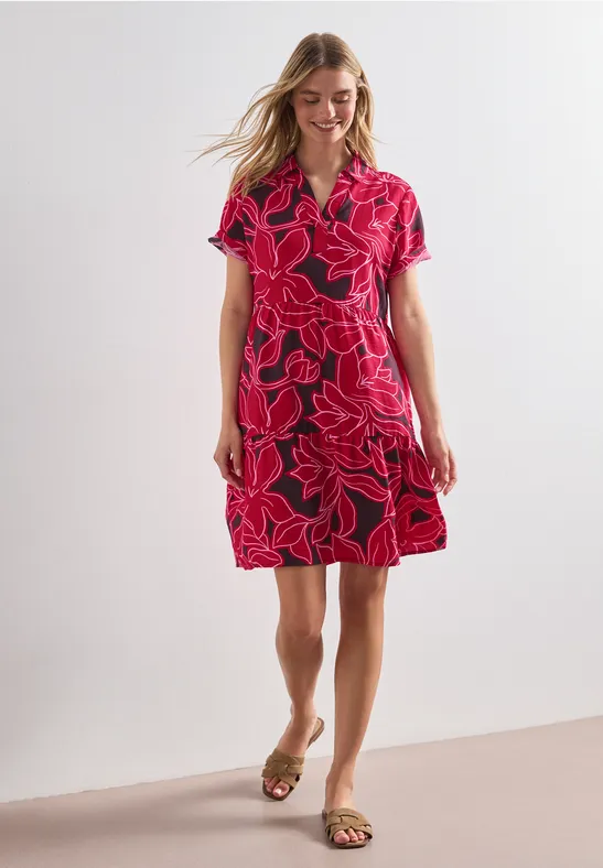 Femme portant une robe rouge mi-longue à motif floral rose, manches courtes et sandales, posant devant un fond clair.