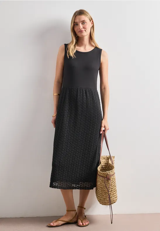 Frau in ärmellosem schwarzem Midikleid mit gehäkeltem Muster, Strohtasche und Sandalen vor weißem Hintergrund.