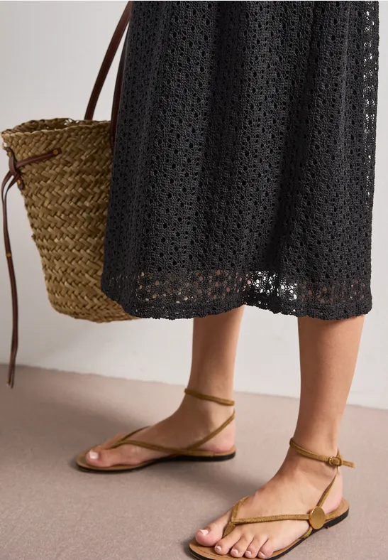 Frau in schwarzem Häkelkleid mit Basttasche und flachen, olivgrünen, geschnürten Sandalen.