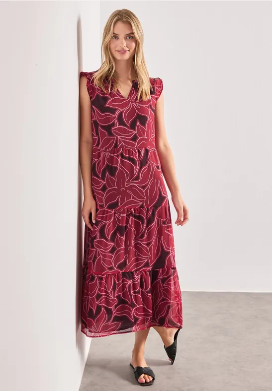 Frau lehnt an Wand, trägt rotes, maximal knöchellanges Kleid mit pinkem Blattmuster und schwarze spitze Ballerinas.