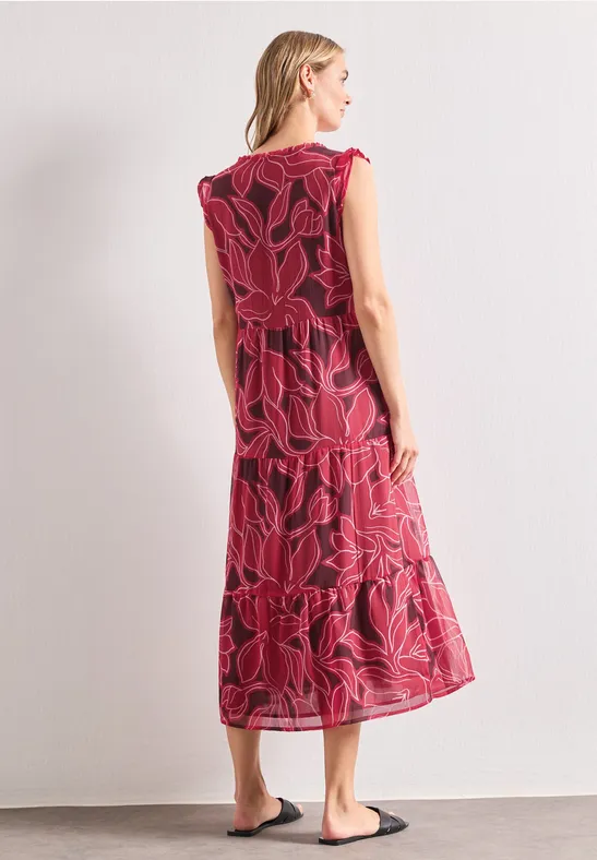 Eine Frau von hinten in einem mittellangen roten Kleid mit großem Blumenmuster, dazu schwarze Sandalen.
