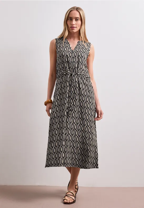 Vrouw in mouwloze, gemêleerde maxi-jurk met ceintuur en sandalen.