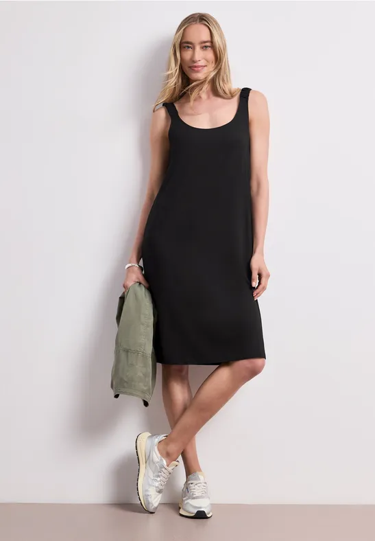 Person trägt ärmelloses schwarzes Kleid, weiße Sneaker und hält eine grüne Jacke.
