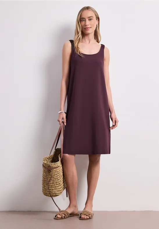 Frau trägt ärmelloses, locker sitzendes lila Kleid, hält Strohtasche und Sandalen.