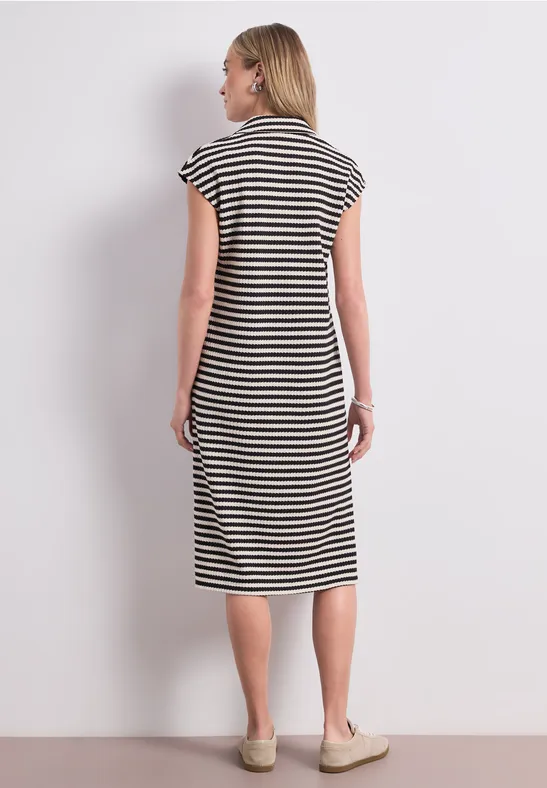 Frau steht rücklings vor weißer Wand, trägt schwarz-weiß gestreiftes Midikleid und beige Schuhe.