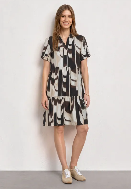 Vrouw in knielange zwart-wit jurk met abstract patroon en witte sneakers, tegen een lichte achtergrond.