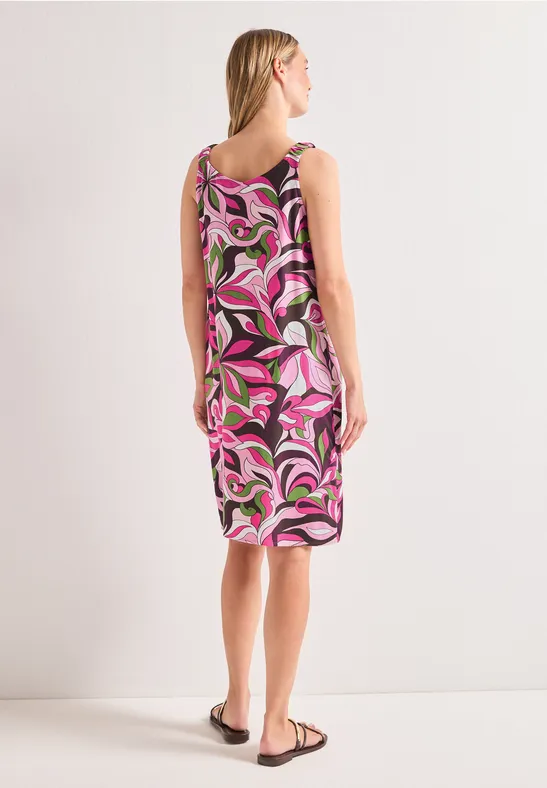 Femme de dos en robe midi sans manches à motif rose, blanc et noir, portant des sandales sur fond clair.
