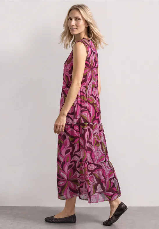 Frau im seitlichen Profil trägt langes ärmelloses Maxikleid mit pink-lila Blumenmuster und flachen Schuhen.