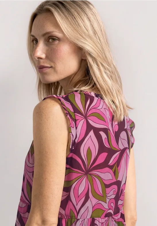 Frau von hinten in ärmelloser Bluse mit pink-lila Blumenmuster, schulterlanges blondes Haar.