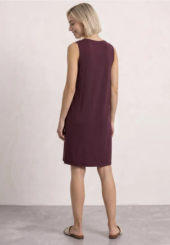 Frau mit kurzen blonden Haaren trägt ärmelloses, knieumspielendes weinrotes Kleid, Ansicht von hinten.