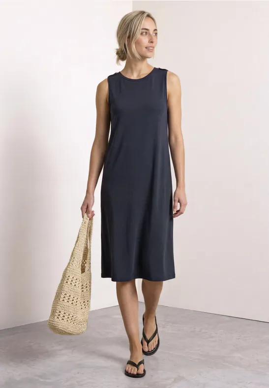 Frau in ärmellosem schwarzem Midikleid mit Strohtasche und Sandalen in neutralem Umfeld.