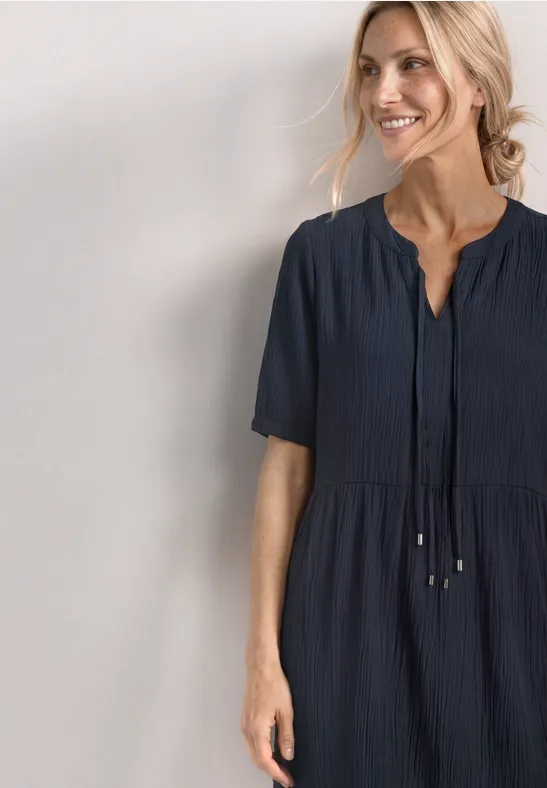 Vrouw in marineblauwe, knielange jurk met korte mouwen en een koord vooraan, zijwaarts gezicht.