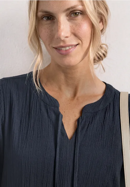 Vrouw in donkerblauw textiel bovenstuk met V-hals, blonde haren nonchalant in een knot.