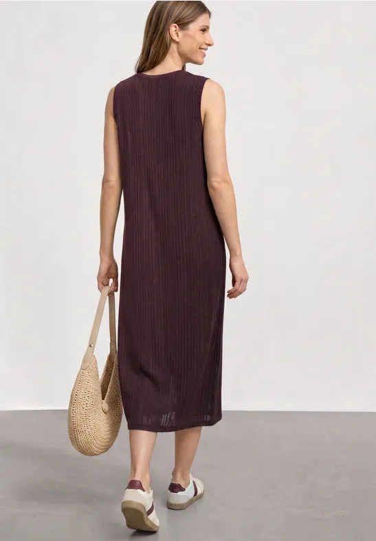 Frau von hinten in ärmellosem, midilangen, dunkelrotem Kleid, mit Strohtasche und weißen Plateauschuhen.