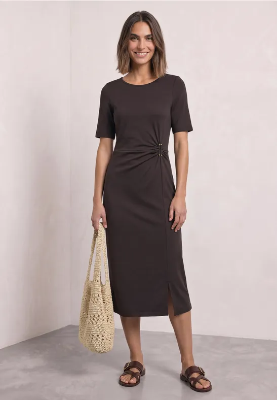 Femme porte robe midi brune cintrée avec fente latérale, sac en crochet écru et sandales plates.