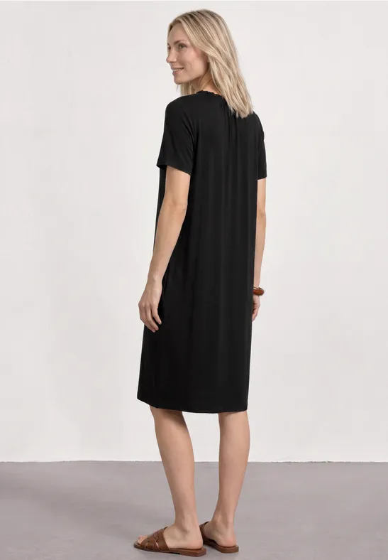Vrouw met blond haar in een zwart midi-jurkje en sandalen, van achteren voor een witte muur.