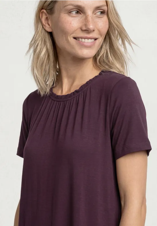Vrouw met halflang blond haar draagt een losse paarse t-shirt tegen een lichte achtergrond