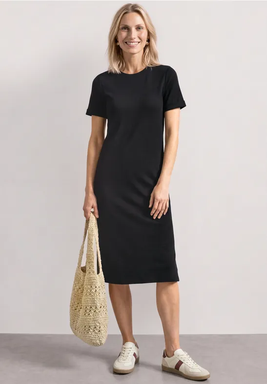Frau in schlichtem schwarzem T-Shirt-Kleid, weiße Sneaker und Netztasche, vor schlichtem Hintergrund.