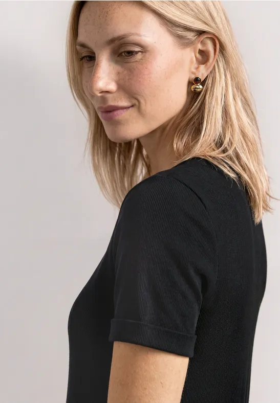Blonde Frau schaut seitlich zur Kamera, trägt schwarzes Kurzarmshirt und goldene Ohrringe.