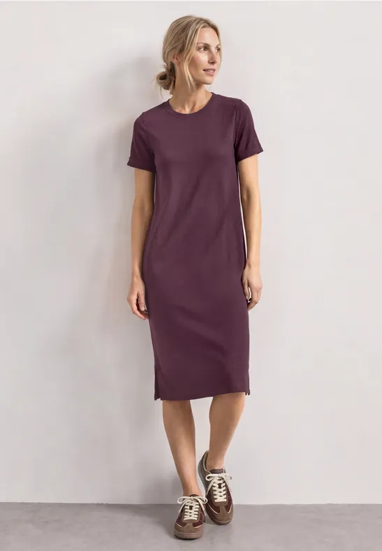Frau trägt schlichtes violettes T-Shirt-Kleid bis zur Wade und Sneaker vor neutralem Hintergrund.