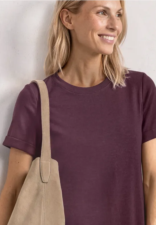 Frau mit blondem Haar trägt ein lila T-Shirt und eine beige Tasche.