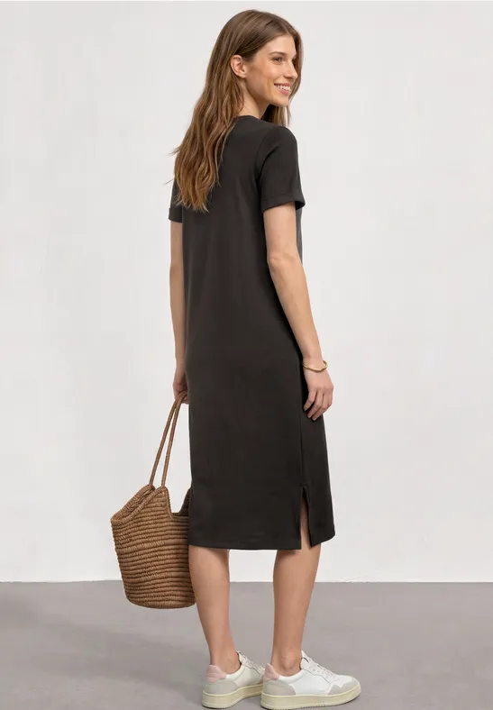 Frau von hinten in knielangem, schwarzem T-Shirt-Kleid und weißen Sneakers, mit Korbtasche