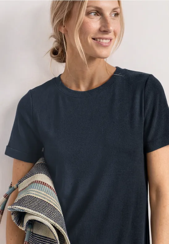 Frau mit dunkelblauem T-Shirt, seitliche Pose, hält gemusterten Stoff unter dem Arm.