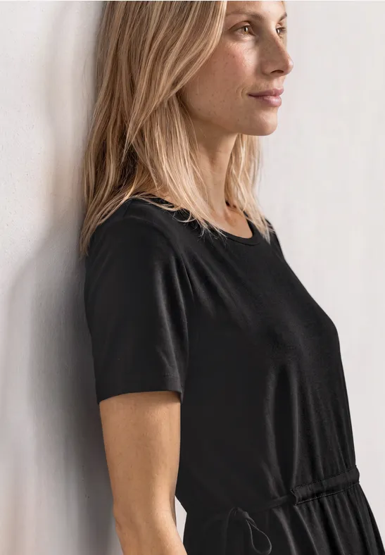 Femme de profil en robe noire à manches courtes adossée à un mur, regard tourné vers la droite.