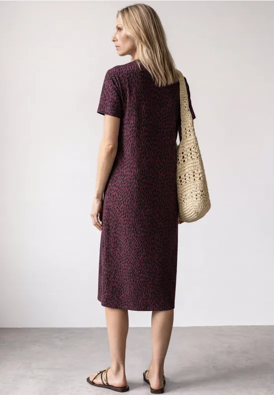 Frau im gemusterten Midi-Kleid, trägt Basttasche und flache Sandalen, steht seitlich vor hellem Hintergrund.