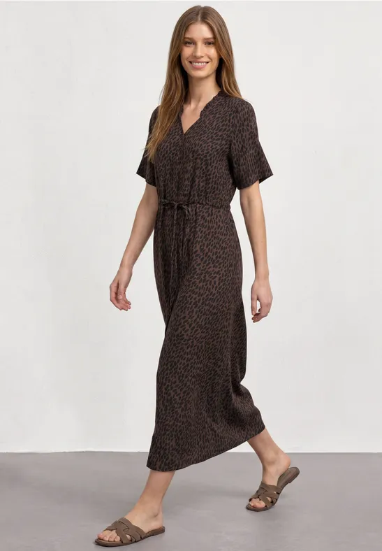 Vrouw in een bruine maxi-jurk met ceintuur en sandalen, die glimlachend loopt.