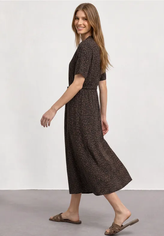 Vrouw in een bruin midi-jurk met patroon en sandalen, glimlachend terwijl ze omkijkt.