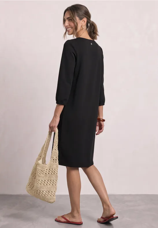 Femme de dos en robe noire genouillère à manches 3/4, porte un sac en macramé et des tongs rouges.
