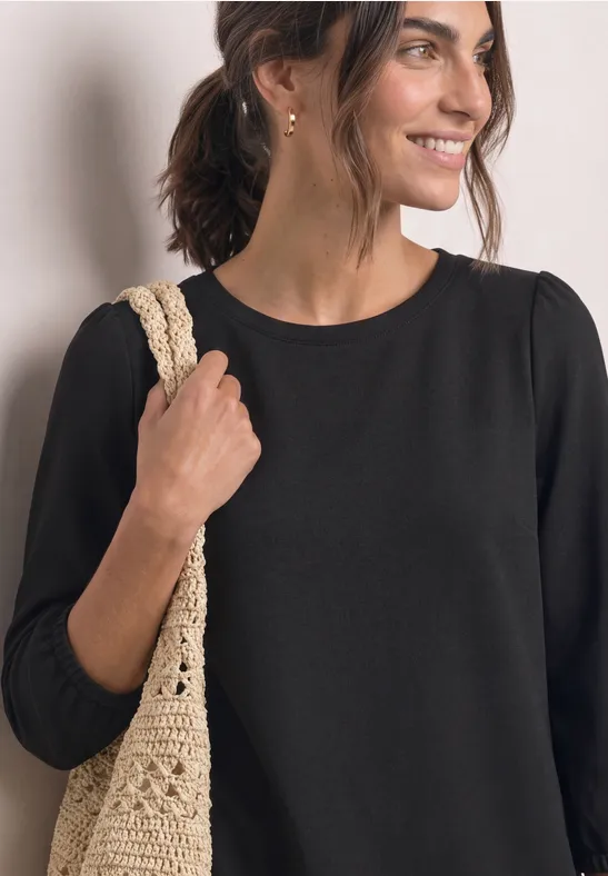 Femme souriante en haut noir, porte un sac beige en crochet sur l’épaule.