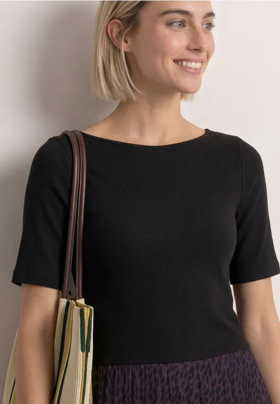 Eine lächelnde Frau in schwarzem Shirt und gemusterter Rock mit Schultertasche, an eine Wand gelehnt.
